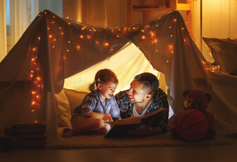 Padre felice della famiglia e figlia del bambino che legge un libro in tenda fotografia stock Padre felice della famiglia e figlia del bambino che legge un libro in tenda fotografia stock