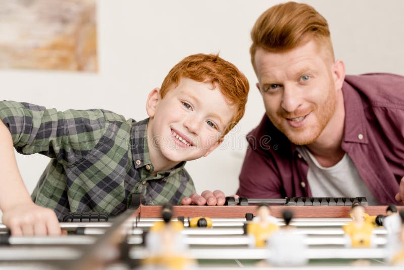 Padre feliz e hijo del pelirrojo que sonríen en la cámara mientras que juega al fútbol de la tabla junto en casa fotografía de archivo libre de regalías