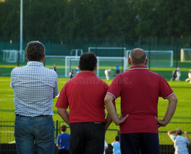 Three fathers standing outside football pitch and watching their sons playing. Three fathers standing outside football pitch and watching their sons playing