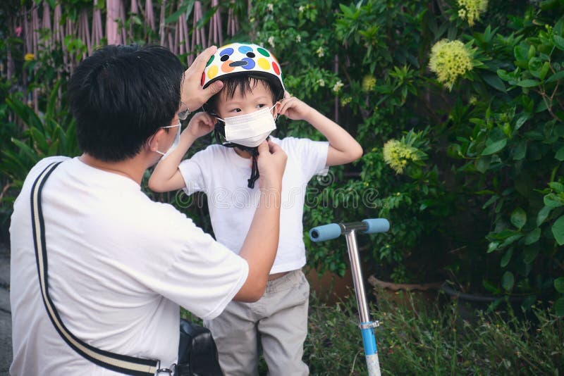 Asian father spending time with little son riding scooter in city, Parent putting mask on his toddler boy child during covid-19 outbreak, New normal lifestyle concept, Selective focus. Asian father spending time with little son riding scooter in city, Parent putting mask on his toddler boy child during covid-19 outbreak, New normal lifestyle concept, Selective focus