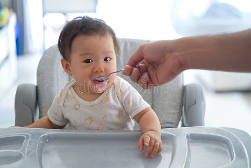 Pai Asiático Que Dá O Papa De Aveia A Sua Criança No Assento De Alimentação Do Bebê Imagem De