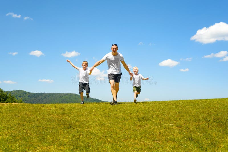 Pai com as duas jovens crianças que correm em conjunto no campo verde em um fundo do céu azul e das nuvens Paternidade e fotografia de stock royalty free Pai com as duas jovens crianças que correm em conjunto no campo verde em um fundo do céu azul e das nuvens Paternidade e fotografia de stock royalty free