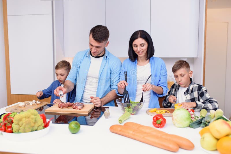 Família jovem e elegante com dois filhos preparando um café da manhã  vegetariano saudável com legumes frescos na cozinha