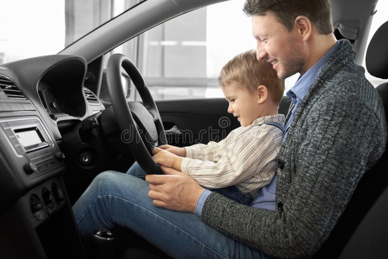 Father holding son on knees, men sitting in driver`s seat of new expensive car. Parent showing to kid car cabin, steering wheel. Happy family observing automobile, smiling. Father holding son on knees, men sitting in driver`s seat of new expensive car. Parent showing to kid car cabin, steering wheel. Happy family observing automobile, smiling.