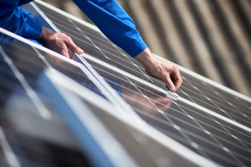 Male engineer in blue suit installing solar photovoltaic panel system. Close up view of electrician hands mounting blue solar module on roof of modern house. Alternative energy ecological concept. Male engineer in blue suit installing solar photovoltaic panel system. Close up view of electrician hands mounting blue solar module on roof of modern house. Alternative energy ecological concept.