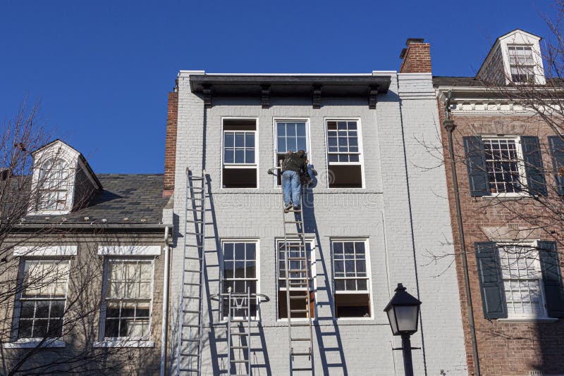 A painter is on top of an industrial size aluminum extension ladder painting the exterior surface of an old brick house.