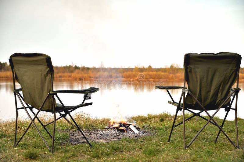 A pair of tourist armchairs, a folding chair, a bonfire on the river bank in the evening at sunset, twilight in autumn at sunset