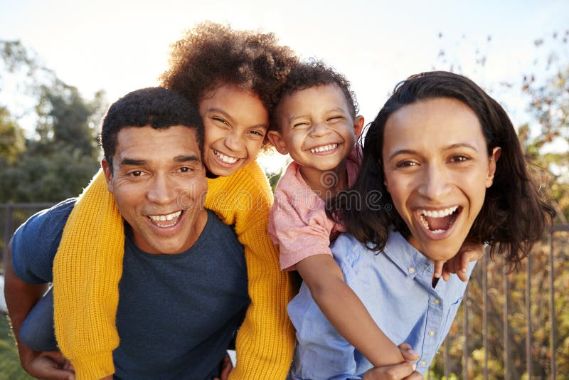 Pais afro-americanos novos dos pais que têm o divertimento que reboca suas crianças no jardim, olhando à câmera e rindo, clos