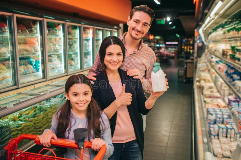 Pais e filha novos na mercearia Estão entre shelfs do produto e pose na câmera Trole da posse da menina imagens de stock Pais e filha novos na mercearia Estão entre shelfs do produto e pose na câmera Trole da posse da menina imagens de stock
