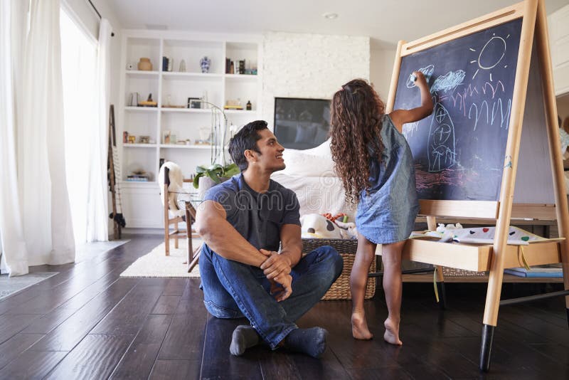 Paizinho latino-americano que senta-se no assoalho na sala de estar que olha seu desenho novo da filha no quadro-negro
