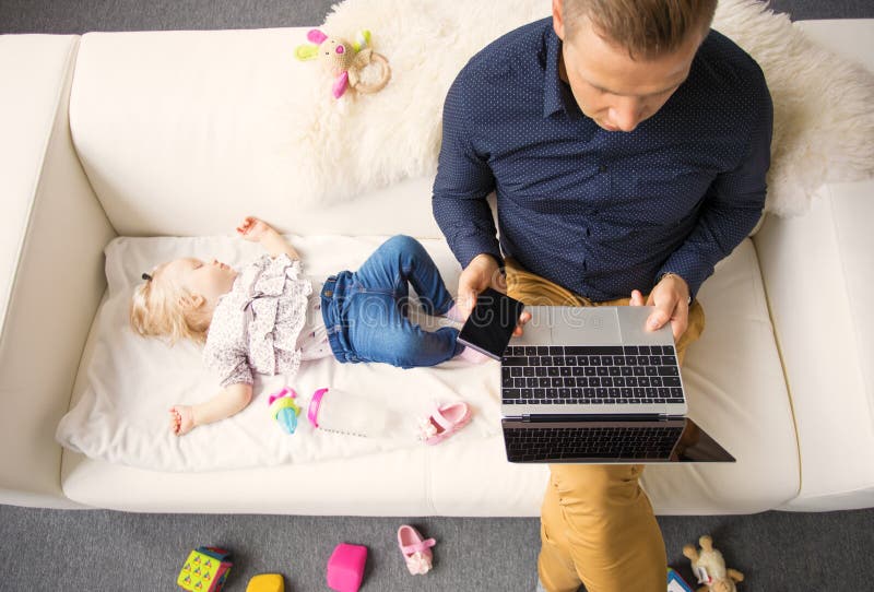Paizinho que trabalha no portátil quando seu bebê que dorme no sofá fotos de stock Paizinho que trabalha no portátil quando seu bebê que dorme no sofá fotos de stock
