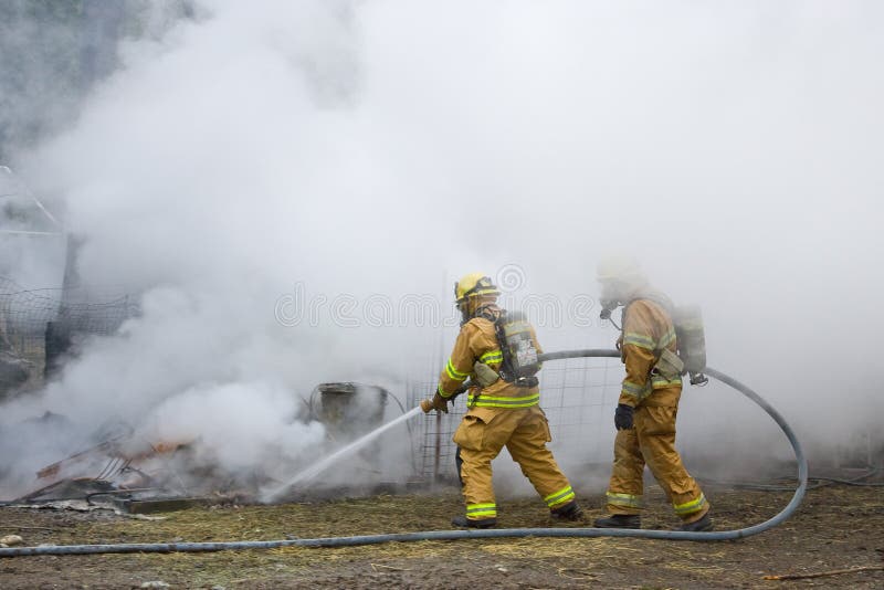 Two firefighters putting hose on outdoor structure fire with smoke and steam background. Two firefighters putting hose on outdoor structure fire with smoke and steam background