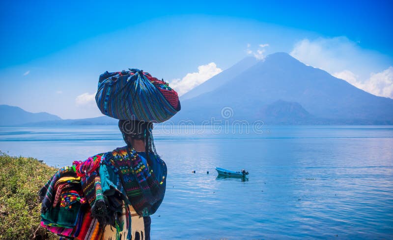 Panajachel, Guatemala -April, 25, 2018: Outdoor view of unidentifed indigenous woman, wearing typical clothes and walking in lakeshore with small boats in Atitlan Lake and volcano in Background in Guatemala. Panajachel, Guatemala -April, 25, 2018: Outdoor view of unidentifed indigenous woman, wearing typical clothes and walking in lakeshore with small boats in Atitlan Lake and volcano in Background in Guatemala.