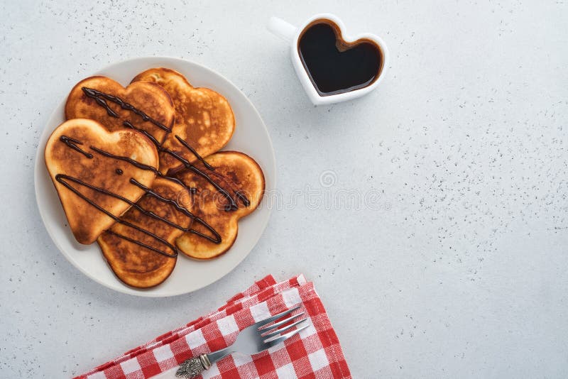 Pancakes in shape of breakfast hearts with chocolate sauce in gray ceramic plate, cup of coffee on gray concrete background. Table