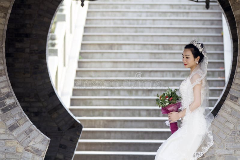 Bride hold bridal bouquet with white wedding dress near a brick arch. wear white crown on her head, black hair, sweet smile, happy. red lip, flower shape earrings, long stairs behind her. vantage building. in shanghai shui bo park, park of water in Shanghai. Bride hold bridal bouquet with white wedding dress near a brick arch. wear white crown on her head, black hair, sweet smile, happy. red lip, flower shape earrings, long stairs behind her. vantage building. in shanghai shui bo park, park of water in Shanghai.