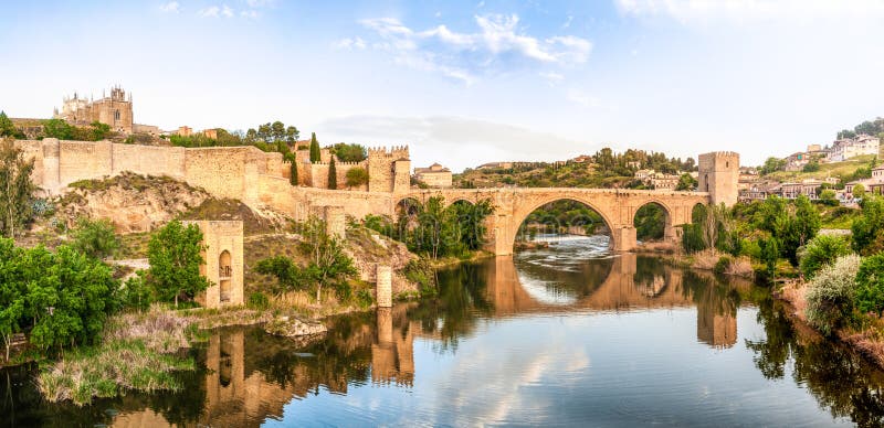 Beautiful landscape of Toledo in Spain. Stone bridge across calm river. Blue sky reflected in crystal clear water. Big fort and country houses in background. Popular tourist place in Europe. Beautiful landscape of Toledo in Spain. Stone bridge across calm river. Blue sky reflected in crystal clear water. Big fort and country houses in background. Popular tourist place in Europe.