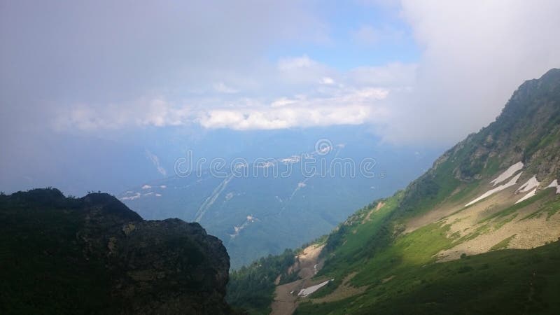 Panorama of the mountains and of the Aibga ridge with low clouds. Remains of snow and fresh green grass on the mountains near the stock photo Panorama of the mountains and of the Aibga ridge with low clouds. Remains of snow and fresh green grass on the mountains near the stock photo