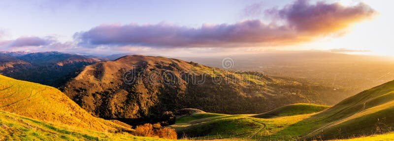 Panoramic sunset view of hills and valleys in the Diablo mountain range starting to turn green; San Jose and bright light from the