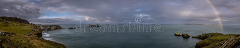 Panoramic view of a rainbow shining between rainy clouds, sea and cliffs near Ballintoy in Northern Ireland, with Sheep Island in