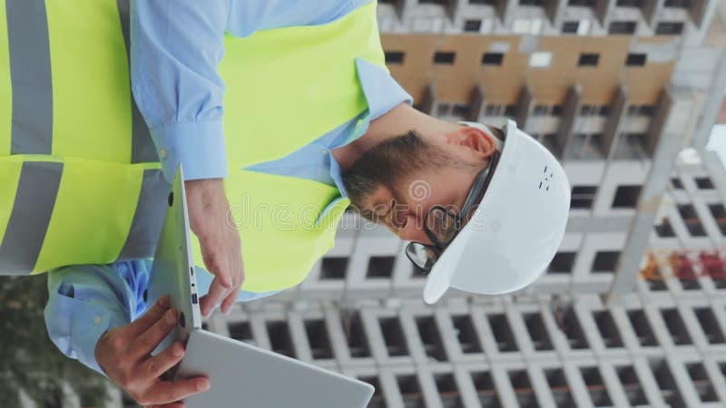 Pantalla vertical : inspector de construcción barbudo haciendo notas sobre el portátil en el sitio de construcción