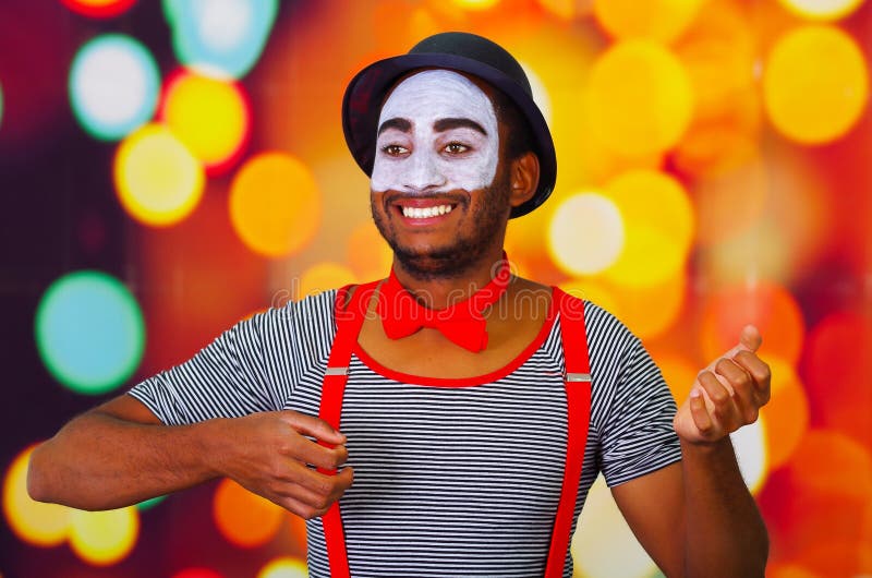 Pantomime man wearing facial paint posing for camera, using hands interacting body language, blurry lights background stock image
