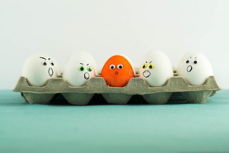 Paper tray full of angry white eggs and only one different orange egg with woman face and long lashes. Discrimination concept. Paper tray full of angry white eggs and only one different orange egg with woman face and long lashes. Discrimination concept.