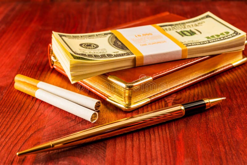 Pack of dollars on a leather diary and cigarettes with golden pen on a mahogany table. Focus on the cigarettes. Pack of dollars on a leather diary and cigarettes with golden pen on a mahogany table. Focus on the cigarettes