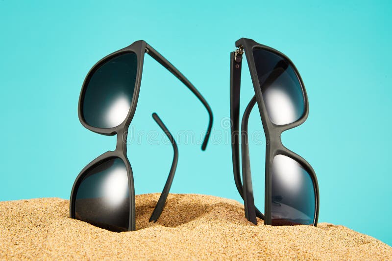 Pair of black sunglasses vertically sticking out of sand on blue background. Side view. Summer vacation on beach, seashore, river coast with stylish sun glasses, shades, eyeglasses. Banner template. Pair of black sunglasses vertically sticking out of sand on blue background. Side view. Summer vacation on beach, seashore, river coast with stylish sun glasses, shades, eyeglasses. Banner template.