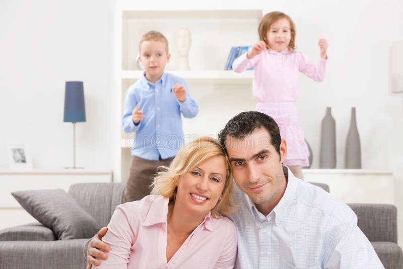 Happy couple sitting at floor at home, embracing. Their children jumping on couch in background. Happy couple sitting at floor at home, embracing. Their children jumping on couch in background.