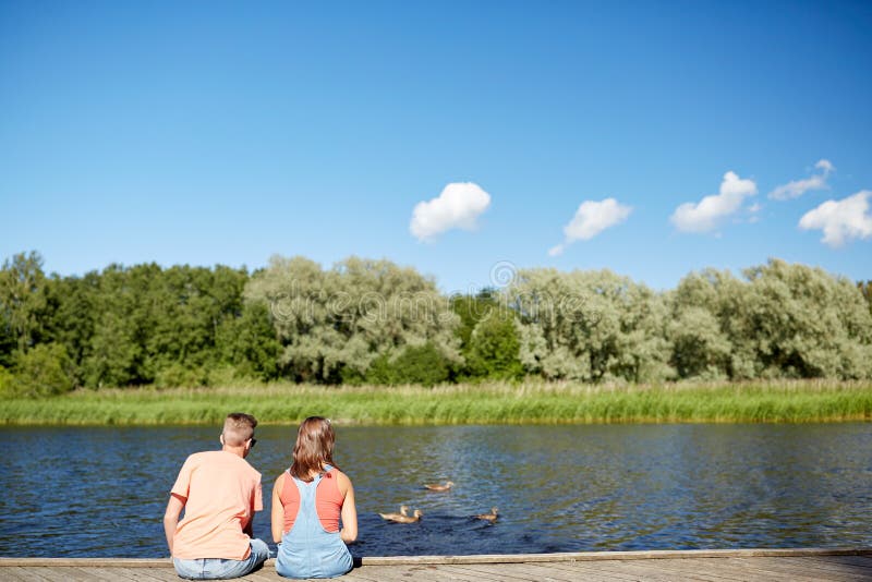 Summer, vacation, love and people concept - happy teenage couple sitting on river berth looking at swimming ducks. Summer, vacation, love and people concept - happy teenage couple sitting on river berth looking at swimming ducks