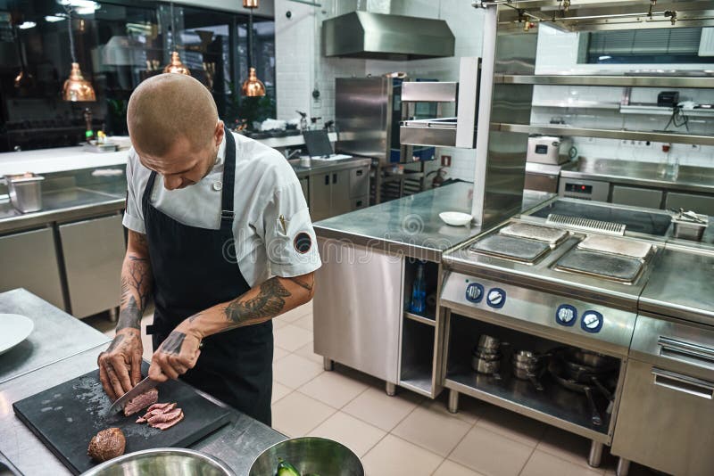 Paraboloïde principal Chef chauve masculin avec de beaux tatouages sur ses mains coupant une viande tout en se tenant dans une cu photographie stock Paraboloïde principal Chef chauve masculin avec de beaux tatouages sur ses mains coupant une viande tout en se tenant dans une cu photographie stock