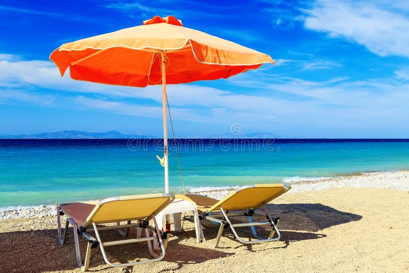 Parapluies de plage colorés avec des chaises de plate-forme Pebble Beach et île dans la distance photographie stock libre de droits Parapluies de plage colorés avec des chaises de plate-forme Pebble Beach et île dans la distance photographie stock libre de droits