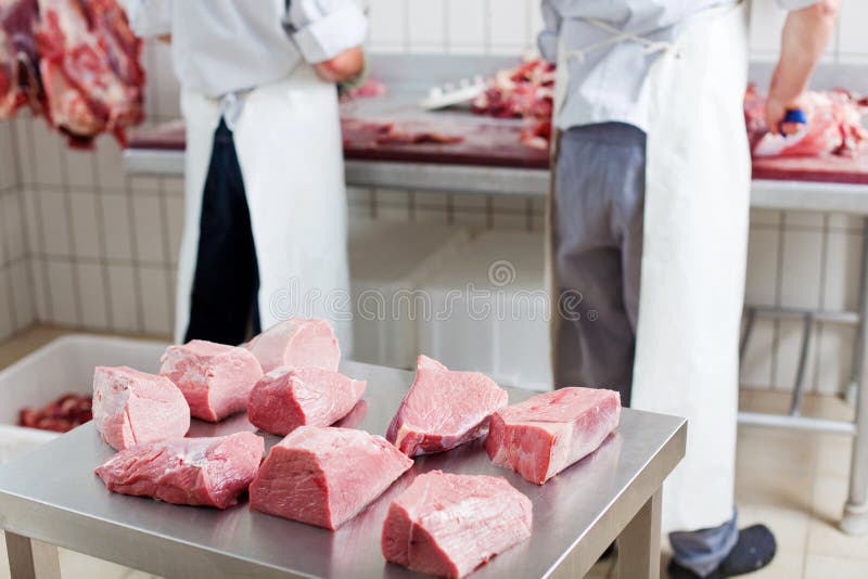 Several portions of fresh cut meat resting on a stainless steel workbench, with two butchers working on the background. Several portions of fresh cut meat resting on a stainless steel workbench, with two butchers working on the background