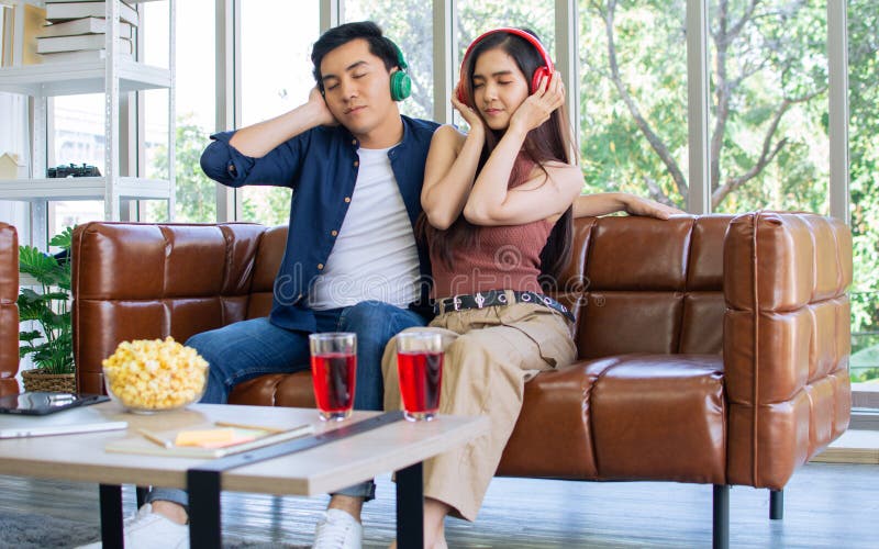 Pareja de asia dulce y adorable sentada en un sofá en la sala de estar en casa usando auriculares escuchando música junto con feli imagen de archivo