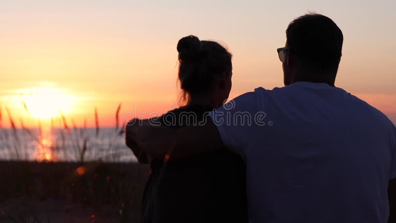 Pareja de jóvenes amantes sentados en la playa en la romántica puesta de sol dorada. vista posterior almacen de video