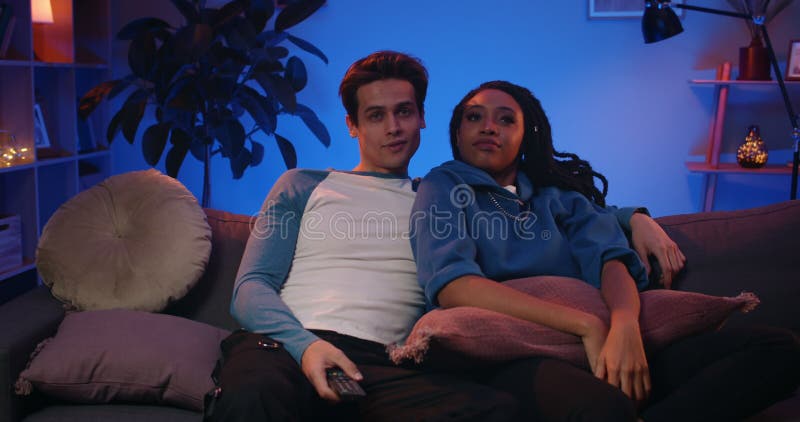 Pareja de jóvenes de etnias mixtas viendo televisión mientras están sentados en el sofá. un tipo guapo girando en tv ang abrazando metrajes