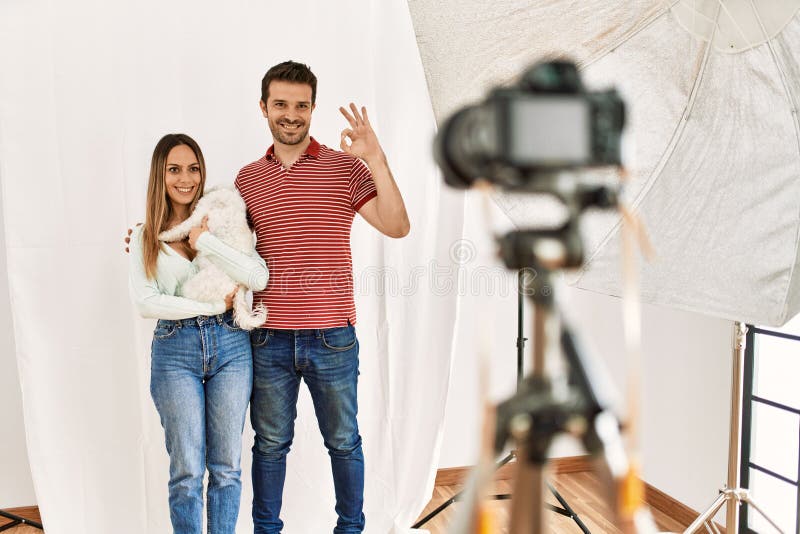 Pareja de novios y novias con perro posando como modelo en el estudio de fotografía haciendo un buen signo con los dedos sonriendo imagenes de archivo