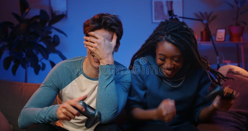 Pareja feliz sosteniendo joysticks inalámbricos y jugando mientras los hombres pierden a la gente millennial disfrutando de un jue almacen de video