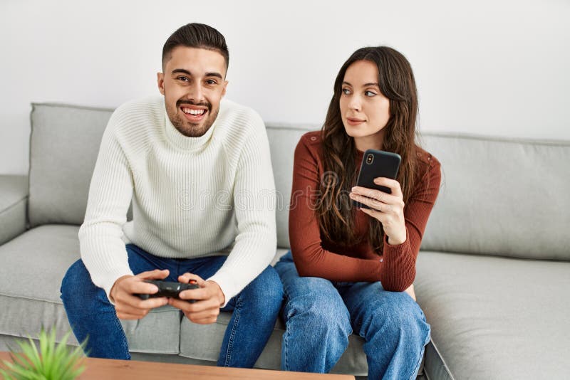 Pareja hispana joven jugando a videojuegos y usando smartphone en casa foto de archivo libre de regalías