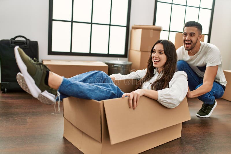 Pareja hispana joven sonriendo feliz jugando usando una caja de cartón como auto en casa nueva fotografía de archivo