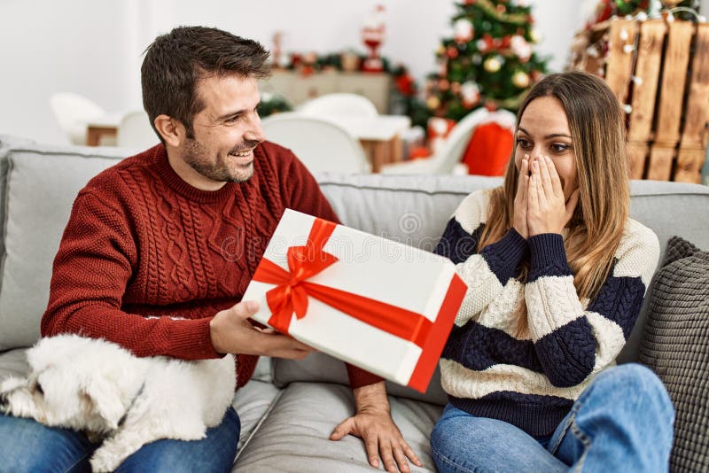 Pareja hispana joven sosteniendo un regalo sentado en el sofá con un perro en casa imágenes de archivo libres de regalías