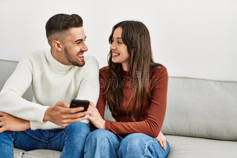 Pareja hispana joven usando un smartphone sentado en el sofá de casa fotos de archivo