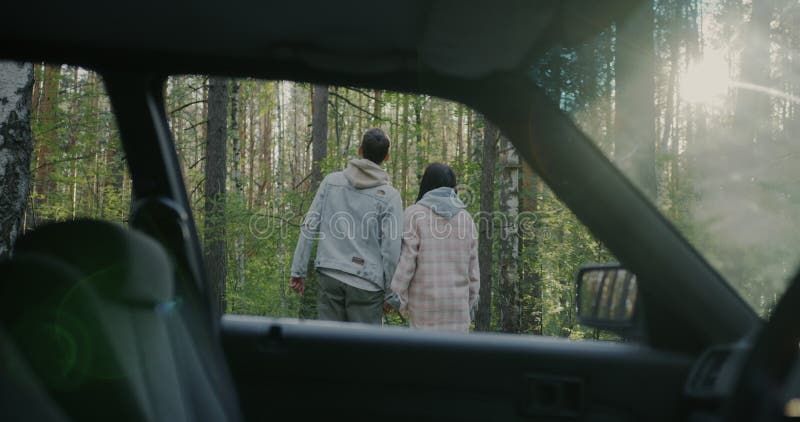 Pareja joven alegre viendo el bosque iluminado por el sol parado cerca de un auto durante un viaje por carretera en el campo