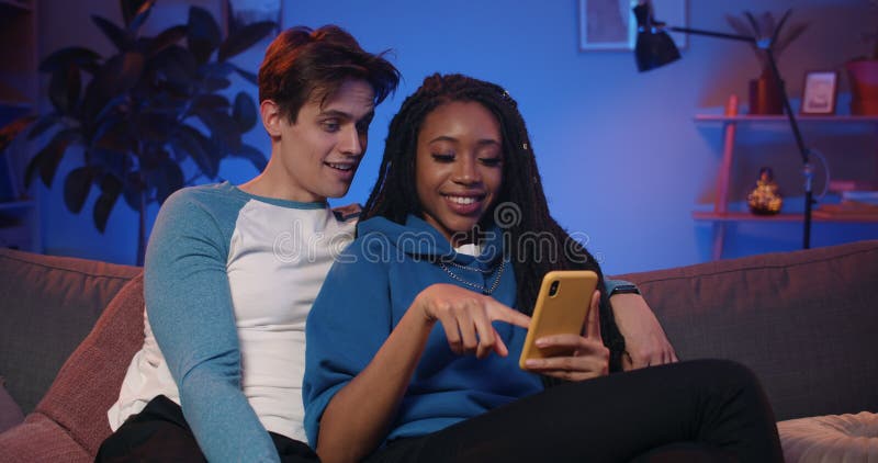 Pareja joven mirando el smartphone y usando aplicaciones divertidas mientras pasaba tiempo juntos en el sofá. sonriente metrajes