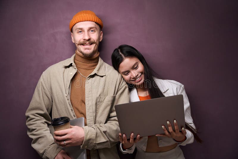 Pareja joven posando con cámara con laptops en las manos. Retrato de mujeres sonrientes y un hombre feliz con computadoras portátiles parados en la pared