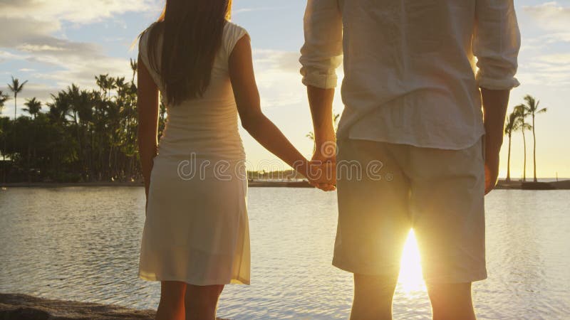 Pareja romántica al atardecer tomándose de la mano en la playa viendo la puesta de sol luna de miel