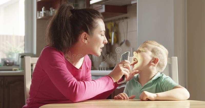Parent Mother is Checking Teeth and Throat of her Child Boy with Mobile Phone Flash Light Sitting at the Table with Sun Shining