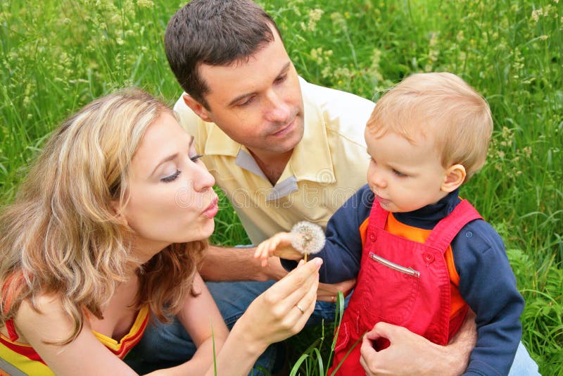 Parents with child sit in grass and blow on dandelion