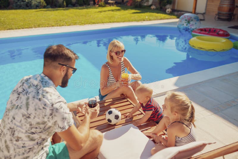 Beauitful young family spending hot sunny summer day by the swimming pool, parents relaxing and drinking cocktails while children are playing on the sun bed next to them. Beauitful young family spending hot sunny summer day by the swimming pool, parents relaxing and drinking cocktails while children are playing on the sun bed next to them