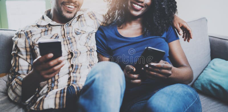Pares afroamericanos felices que se relajan junto en el sofá Hombre negro joven y su novia que usa los teléfonos móviles mientras fotos de archivo libres de regalías Pares afroamericanos felices que se relajan junto en el sofá Hombre negro joven y su novia que usa los teléfonos móviles mientras fotos de archivo libres de regalías